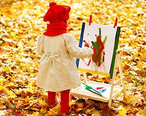 Foto de una niña pintando en un caballete afuera, rodeada de hojas caídas