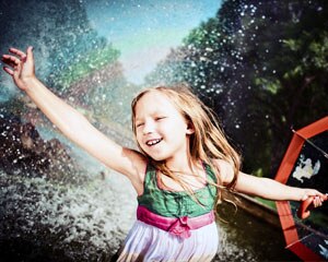 Young girl dances in a water spray that has a rainbow while holding an umbrella